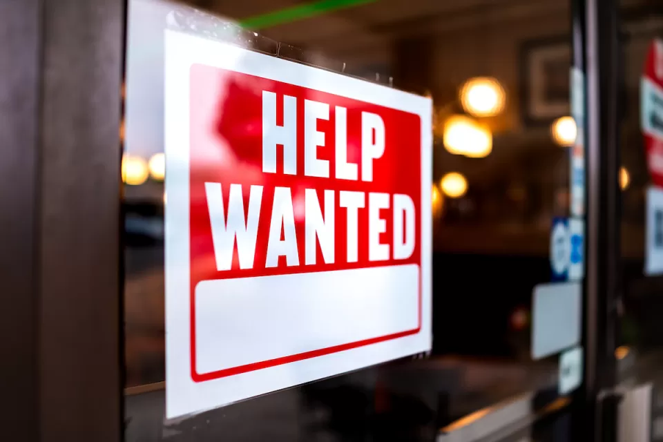 Close-up of Help Wanted sign with white text on red background posted inside storefront window.